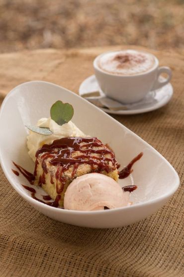 a white bowl containing quandong slice, cream, ice cream and quandong sauce is in the foreground; a cappuccino is in the background