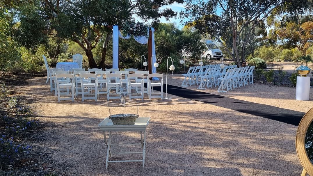 The wedding site consists of a large gravelled area surrounded by native garden. The area is longer than it is wide, with a wooden wedding arbour at one end. The wedding area is currently set up with white cloth and flowers decorating the arbour, rows of white chairs facing the arbour, and a black carpet runner creating an aisle.
